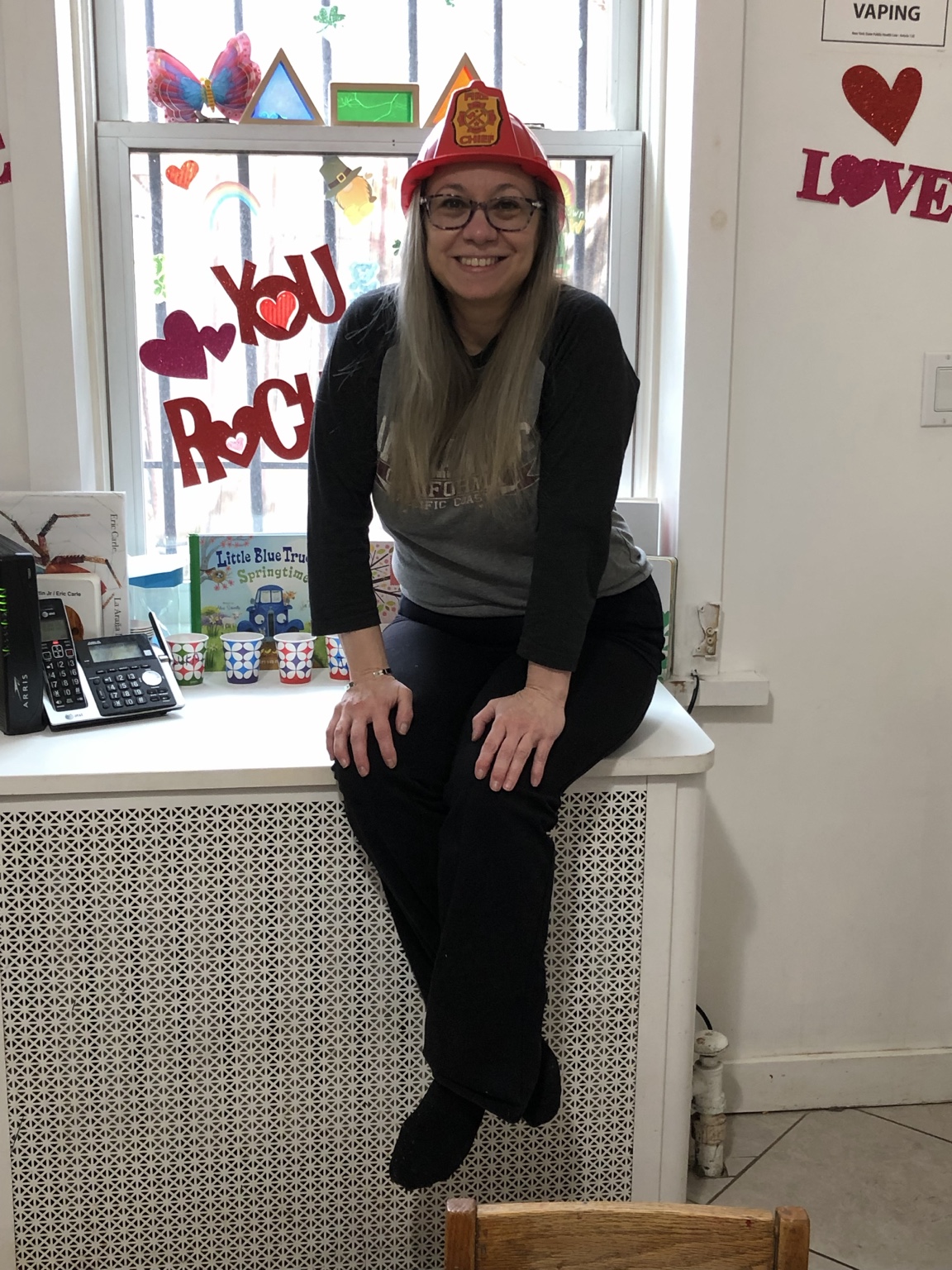Ms. Sandy Roses smiling in a red toy firefighter helmet beside a decorated window at the daycare